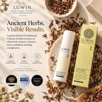 A flat-lay product photograph of LUWIN Ultimate Repair & Clear Face Cream for acne-prone skin, showing the white cream tube and its pale yellow packaging box surrounded by loose dried herbs on a textured beige stone surface.