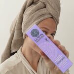 Person holding the packaging of the LUWIN All-rounder Calming & Radiance-Enhancing Facial Mist with an elegant design and soothing purple tones. The model's relaxed post-shower vibe and the sleek packaging suggest a luxurious, spa-like skincare experience.