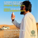 A man in a white robe sprays LUWIN All-Rounder Calming & Radiance-Enhancing Face Mist on his face in the desert, with sand dunes and blue sky in the background. The product is promoted as a summer travel essential for hydration, soothing sensitive skin, reducing redness, brightening complexion, and strengthening the skin barrier.